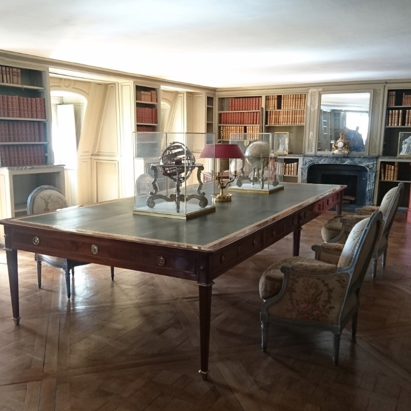 Bibliothekszimmer mit Bücherregalen an allen vier Wänden, einem riesigen Holztisch und zwei Globen, die durch kleine Glasvitrinen geschützt sind.