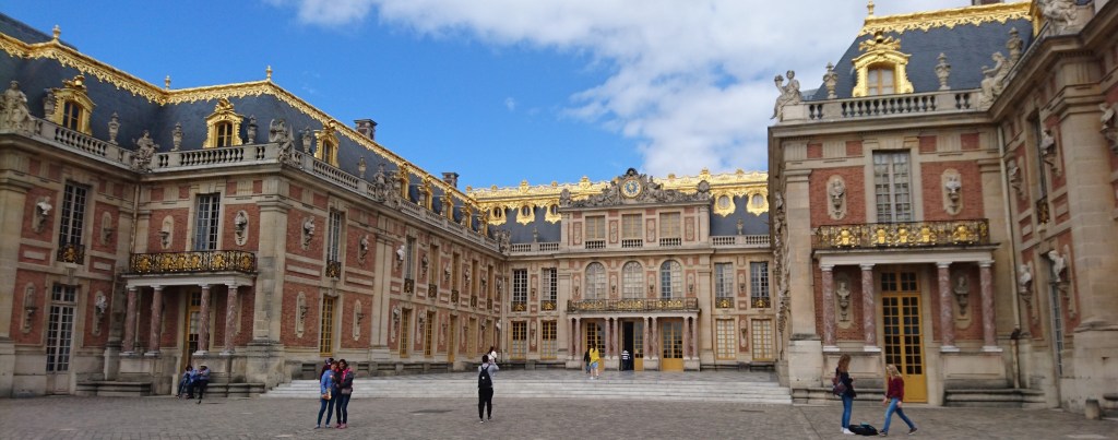 Schlossfassade zur Straße hin, Eingang nach Versailles. Goldene Verzierungen auf dem Dach und roter Stein mit hellen Steinen in einem ausgewogenen Mix.