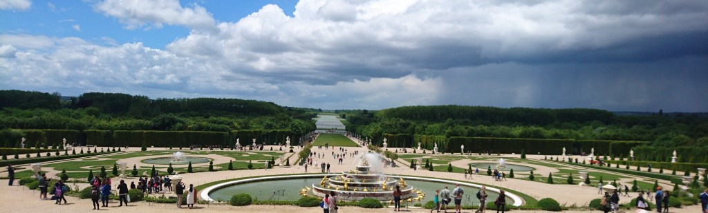 Schlosspark Versailles bei düsterem Wetter. Dicke Wolken hängen über dunklem Grün der Bäume. Der Springbrunnen ist an und Menschen flanieren über die Wege-