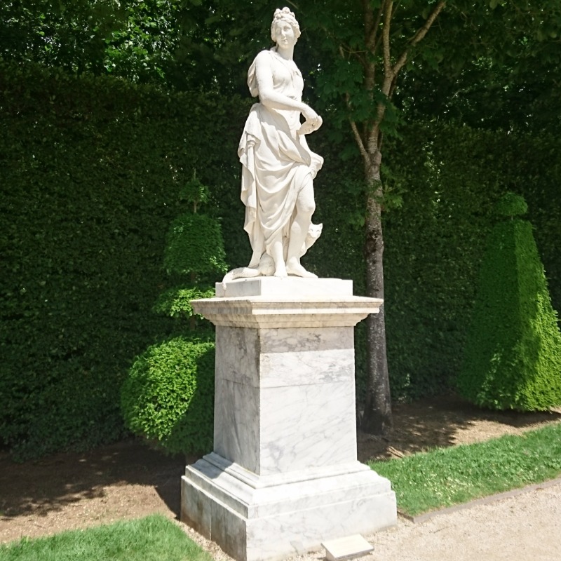Statue einer jungen Frau auf einem Marmorsockel, dahinter eine grüne Hecke.