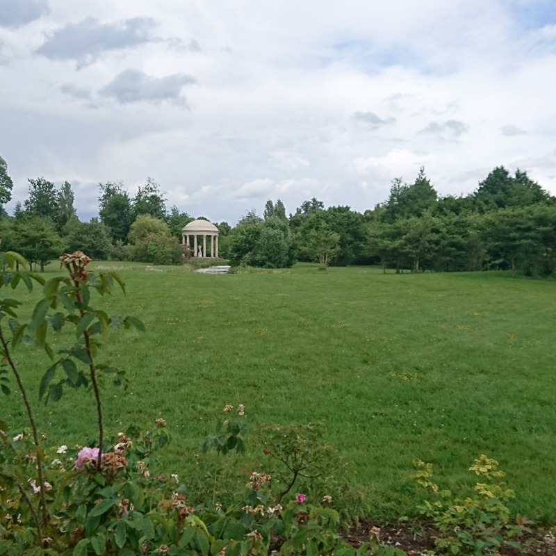 Eine saftig grüne Wiese von Bäumen umsäumt mit einer Hecke und einem kleinen Pavillon im Hintergrund.