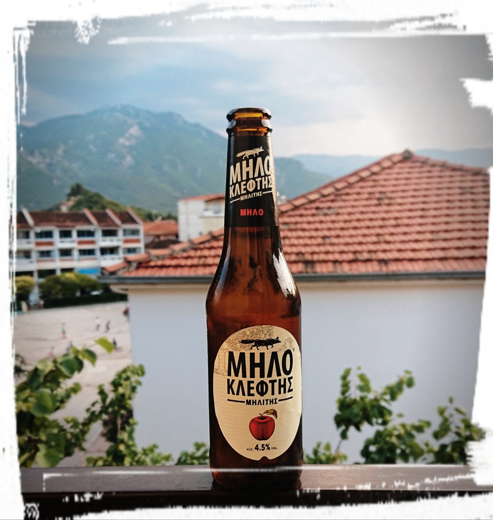 Eine Bierflasche, im Hintergrund ein Haus. Auf der Flasche steht "Milo", das griechische Wort für Apfel, in der Flasche ist Cider.
