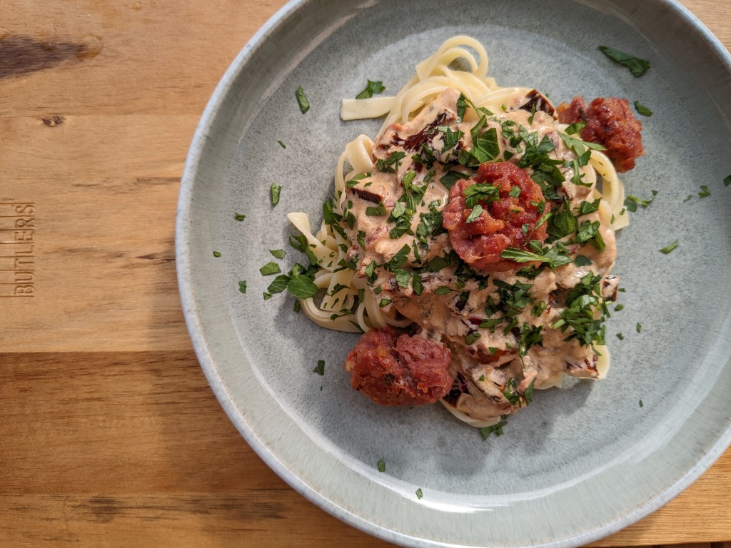 Auf einem runden Teller angerichtete Pasta mit Feta-Sauce mit getrockneten Tomaten, gebratenen Salsiccia-Bällchen und Petersilie.